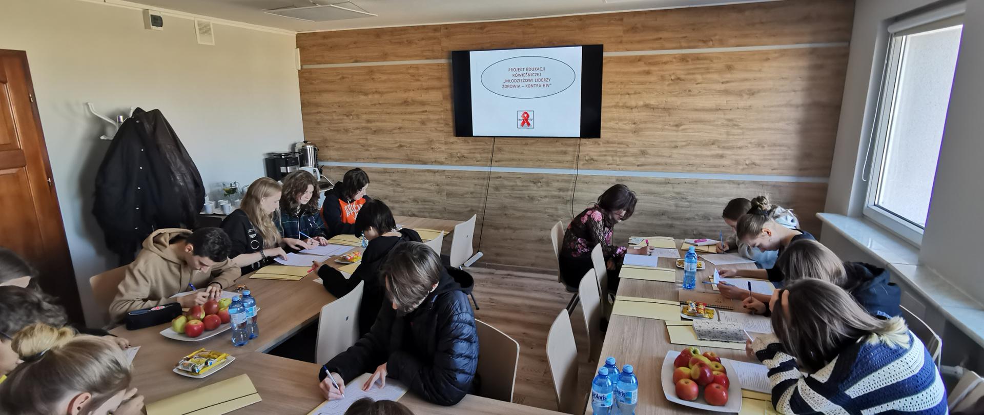 Kilkunastu młodych ludzi siedzi przy stołach w niewielkiej sali konferencyjnej. Na ścianie ekran z wyświetlana prezentacją.