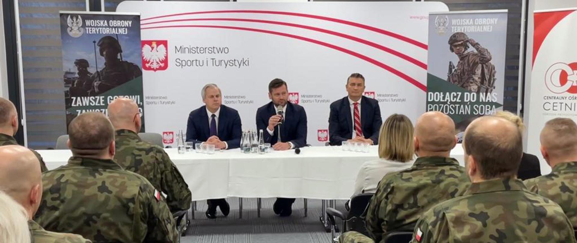 Na pierwszym planie zdjęcia znajduje się widownia złożona z żołnierzy Wojsk Obrony Terytorialnej. Przed nimi, za stołem prezydialnym zasiadło trzech elegancko ubranych mężczyzn. W tle biało-czerwona ścianka wizerunkowa Ministerstwa Sportu i Turystyki oraz materiały wizerunkowe Wojsk Obrony Terytorialnej.