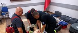 Zdjęcie przedstawia trzech druhów jednostek OSP. Mężczyźni znajdują się w sali Komendy Powiatowej PSP w Mławie. Jeden z mężczyzn wykonuje uciśnięcia klatki piersiowej na manekinie do resuscytacji krążeniowo oddechowej, drugi trzyma maskę resuscytacyjną, a trzeci, w okularach, obserwuje i instruuje. Obok manekina leży defibrylator. W tle widoczne są torby medyczne i polska flaga.