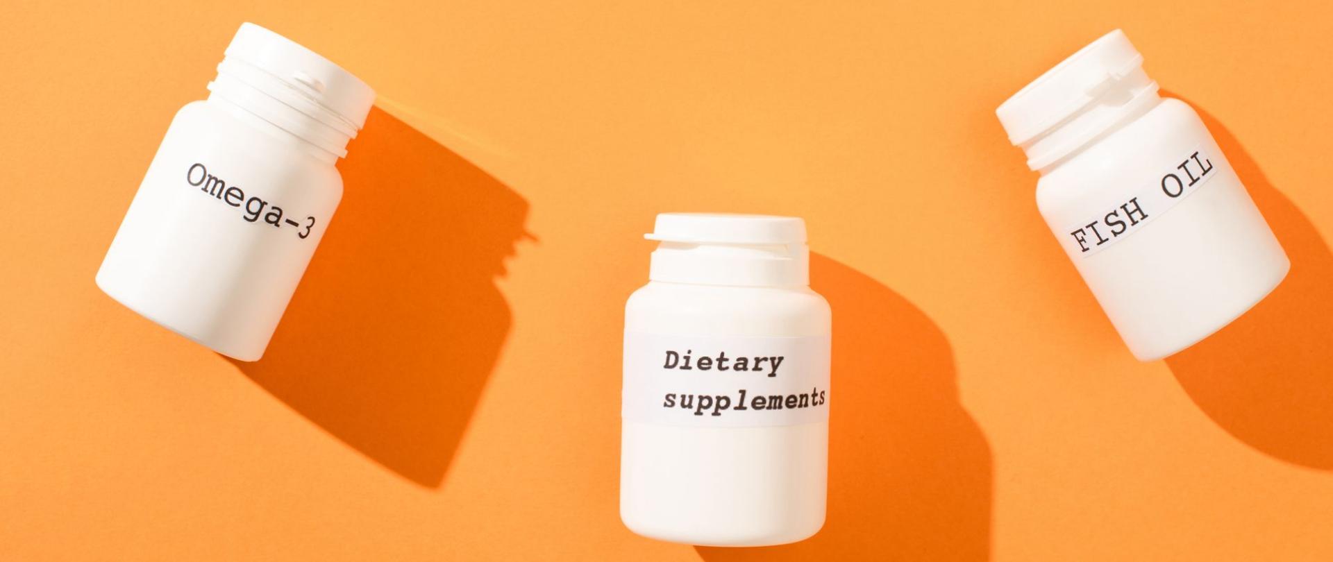 Na zdjęciu są trzy białe pudełka z suplementami diety: omega-3, dietary suplements oraz fish oil. Tło jest pomarańczowe.