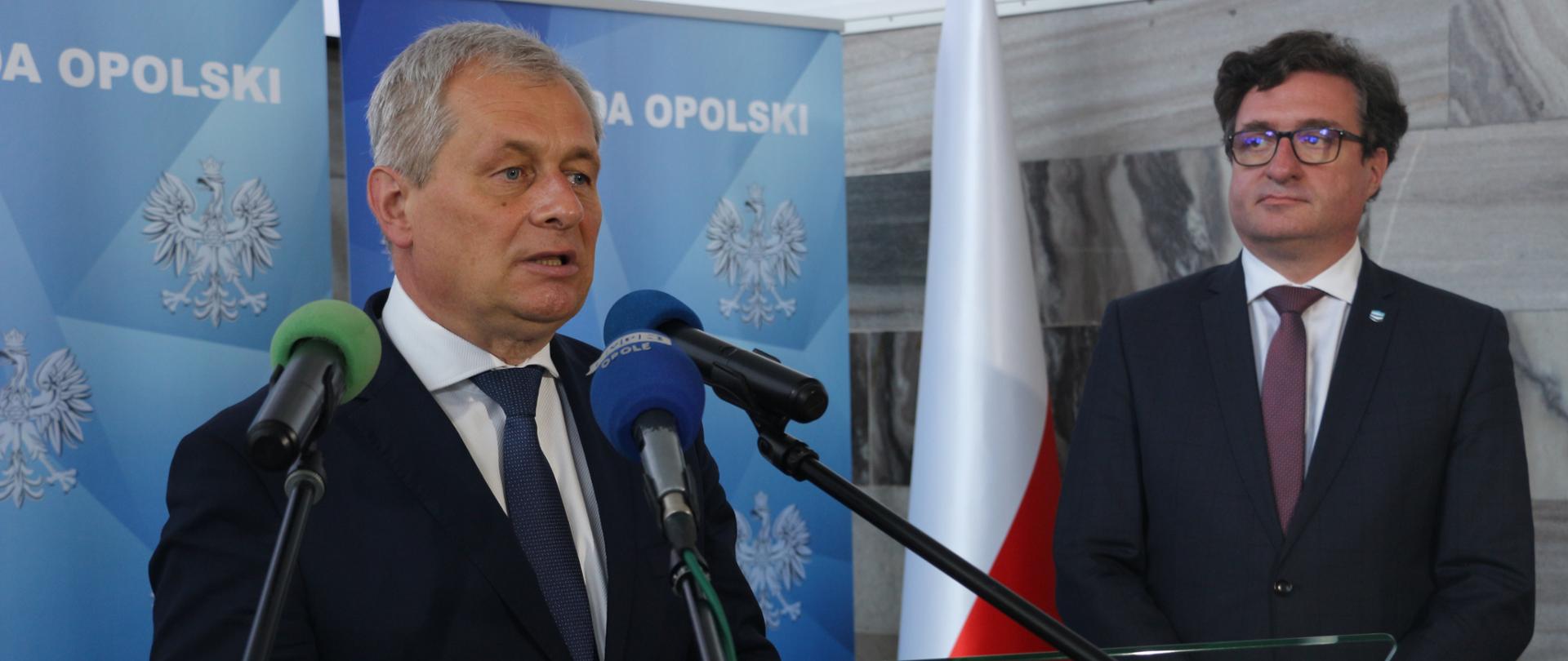 Na zdjęciu na środku kadru stojący za 3 mikrofonami na statywach, wojewoda opolski. Ma czarny garnitur, białą koszulę i granatowy krawat. Przed wojewodą stoi szklana mównica. Wojewoda w dłoniach trzyma okulary korekcyjne. Obok niego - po prawej stronie kadru, stoi wysoki, ciemnowłosy mężczyzna. To prezes PGW Wody Polskie Przemysław Daca. Mężczyzna ustawiony jest frontem do aparatu, spogląda w kierunku wojewody. Za mężczyzna stoi biało-czerwona flaga. Za wojewodę w tle stoi ścianka z napisem Wojewoda Opolski. W dalekim tle szara ściana, marmurowa. 