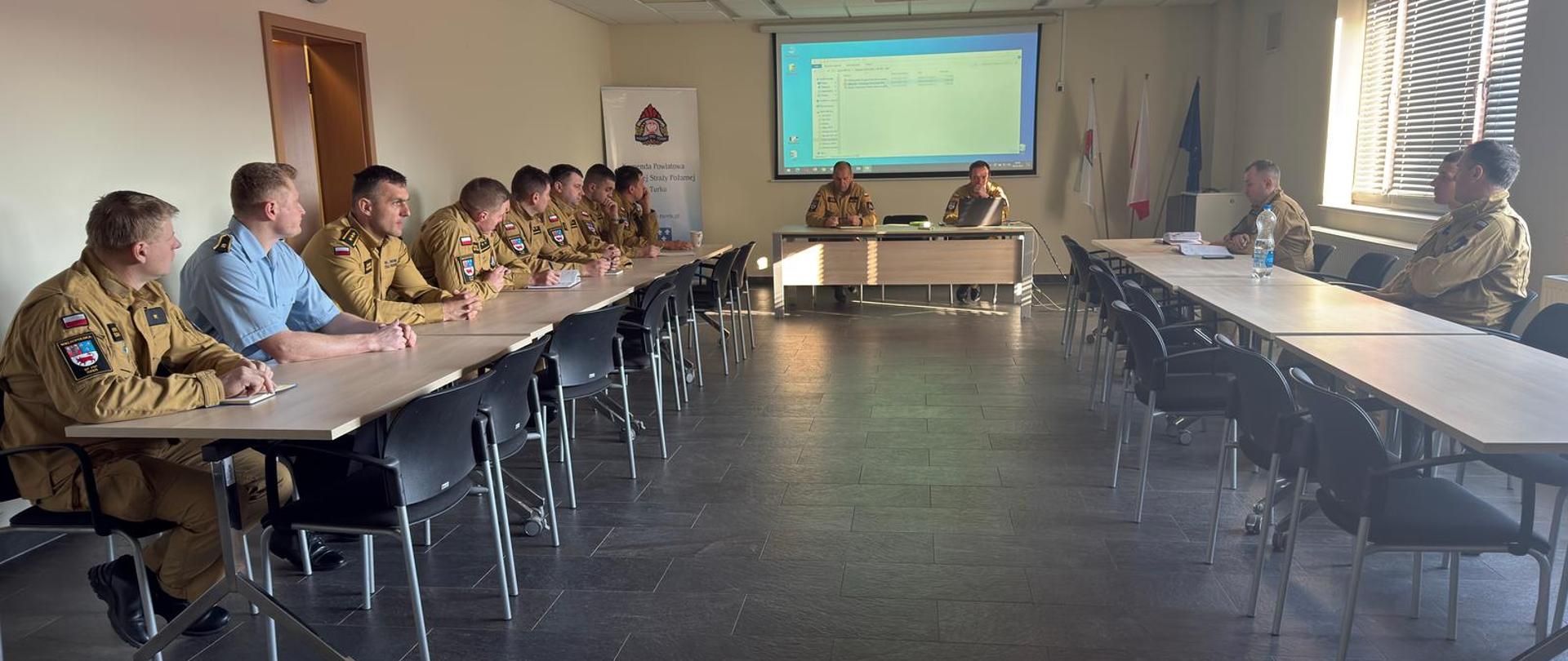 Funkcjonariusze siedzą przy stołach uczestnicząc w odprawie. 