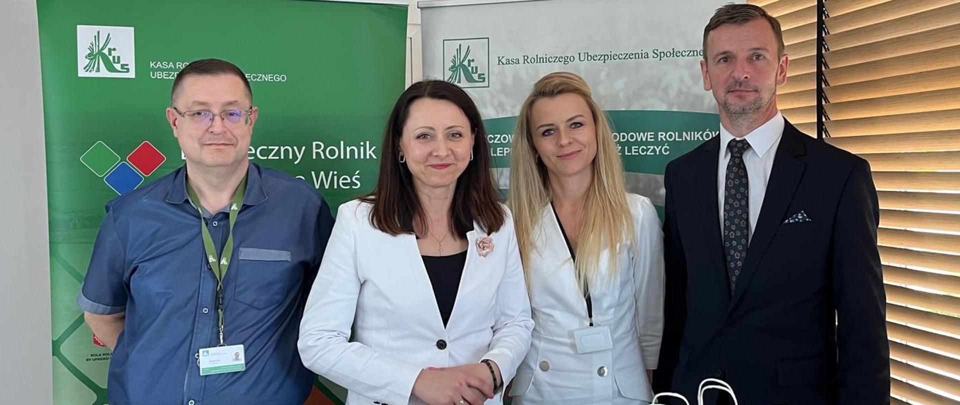 Dwie kobiety i dwóch mężczyzn stoją na tle roll-upów Kasy. Przed nimi znajduje się stoli, na którym są wyłożone materiały prewencyjne Kasy.