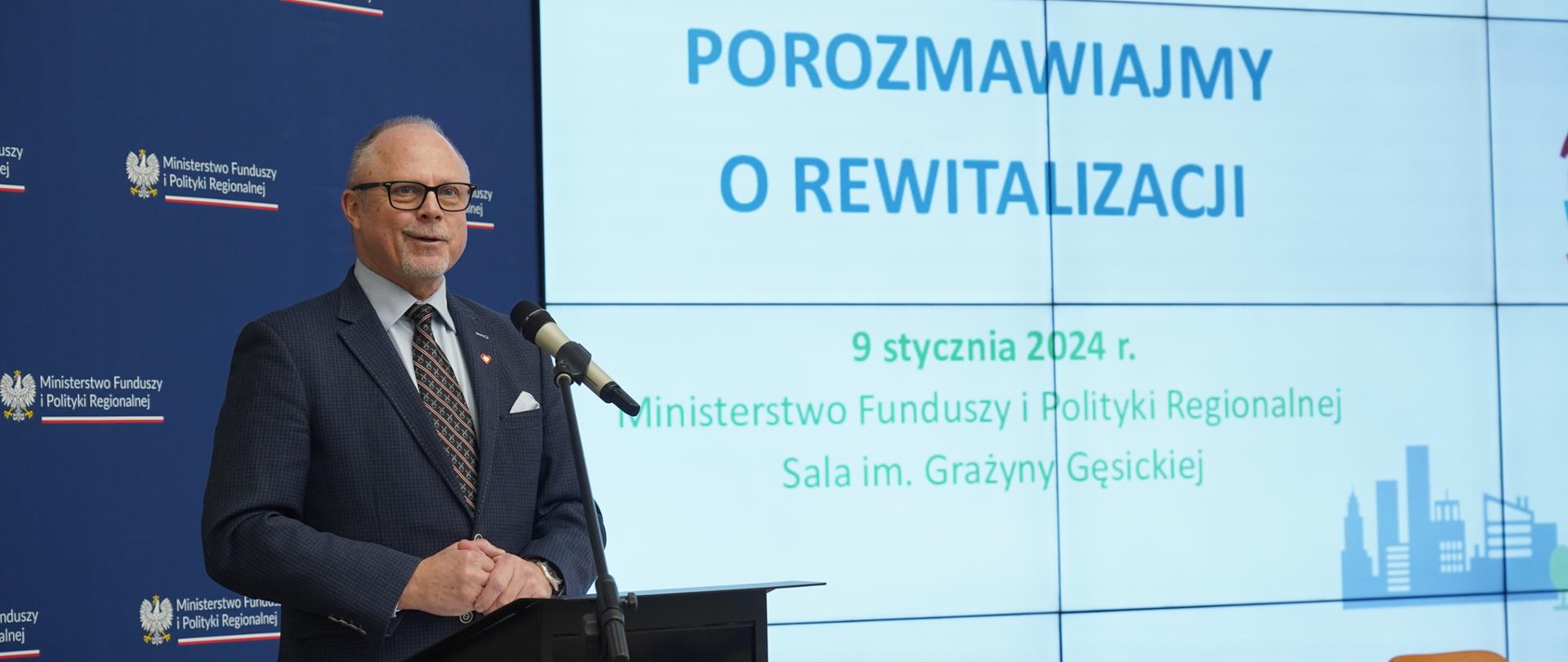 Wiceminister Jacek Protas w mównicy z mikrofonem na tle ścianki MFiPR
