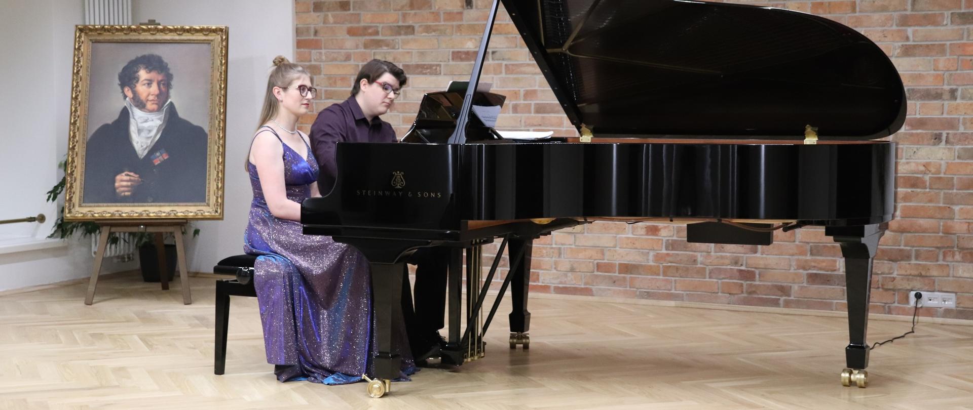Dwie osoby siedzą przy jednym fortepianie, grając na cztery ręce; w tle widoczny jest portret na sztaludze.
