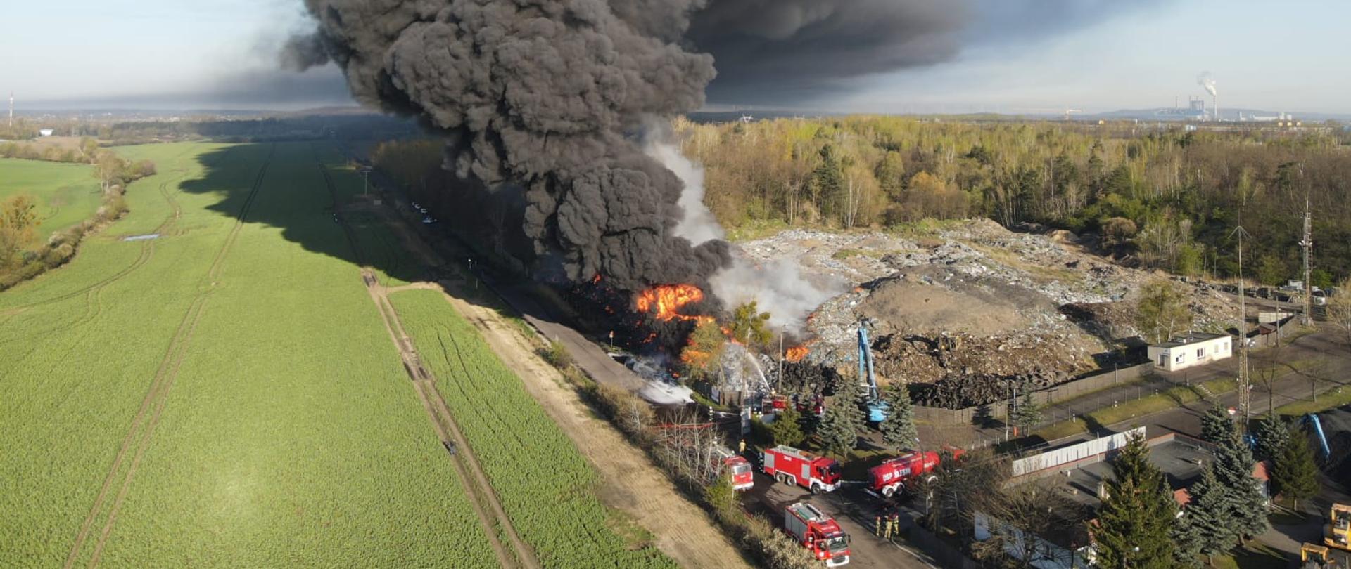 Zdjęcie przedstawia palące się odpady na składowisku odpadów. Gęsty dym unosi się nad składowiskiem. Widać również samochody strażackie.