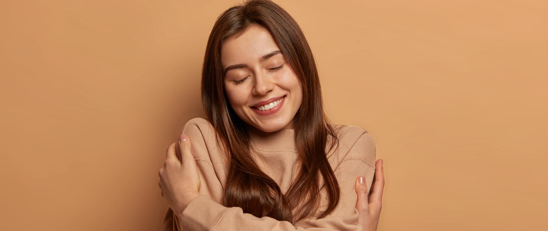 I love myself. Pretty smiling woman embraces herself gently, expresses self love, keeps eyes closed with pleasure, feels comfortable and fullfilled, being egoistic person, isolated on brown background