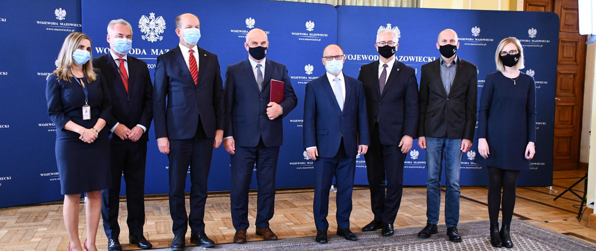 Na zdjęciu znajduje się osiem osób - dwie kobiety i sześciu mężczyzn. Osoby te są elegancko ubrane. Pozują one na tle granatowej ścianki wizerunkowej Wojewody Mazowieckiego.