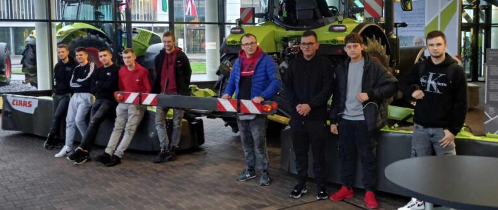 Dziewięciu młodych mężczyzn pozuje do zdjęcia w przeszklonym salonie maszyn rolniczych. Część grupy siedzi na elemencie zielonej maszyny marki Claas, a pozostali stoją obok niej. W tle, za dużymi oknami, widoczne są flagi oraz nowoczesny budynek biurowy.