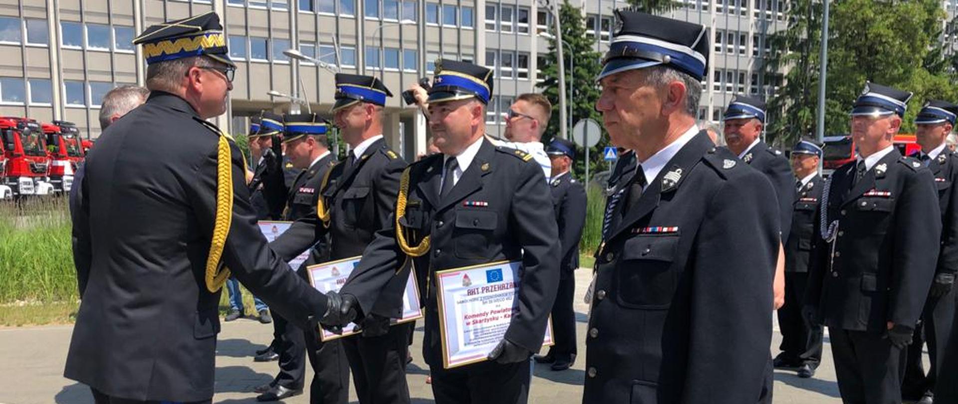 Zastępca Komendanta Głównego PSP wręcza akt przekazania samochodu funkcjonariuszowi PSP, strażacy PSP i OSP ustawieni w rzędach odbierają akty przekazania / promesy