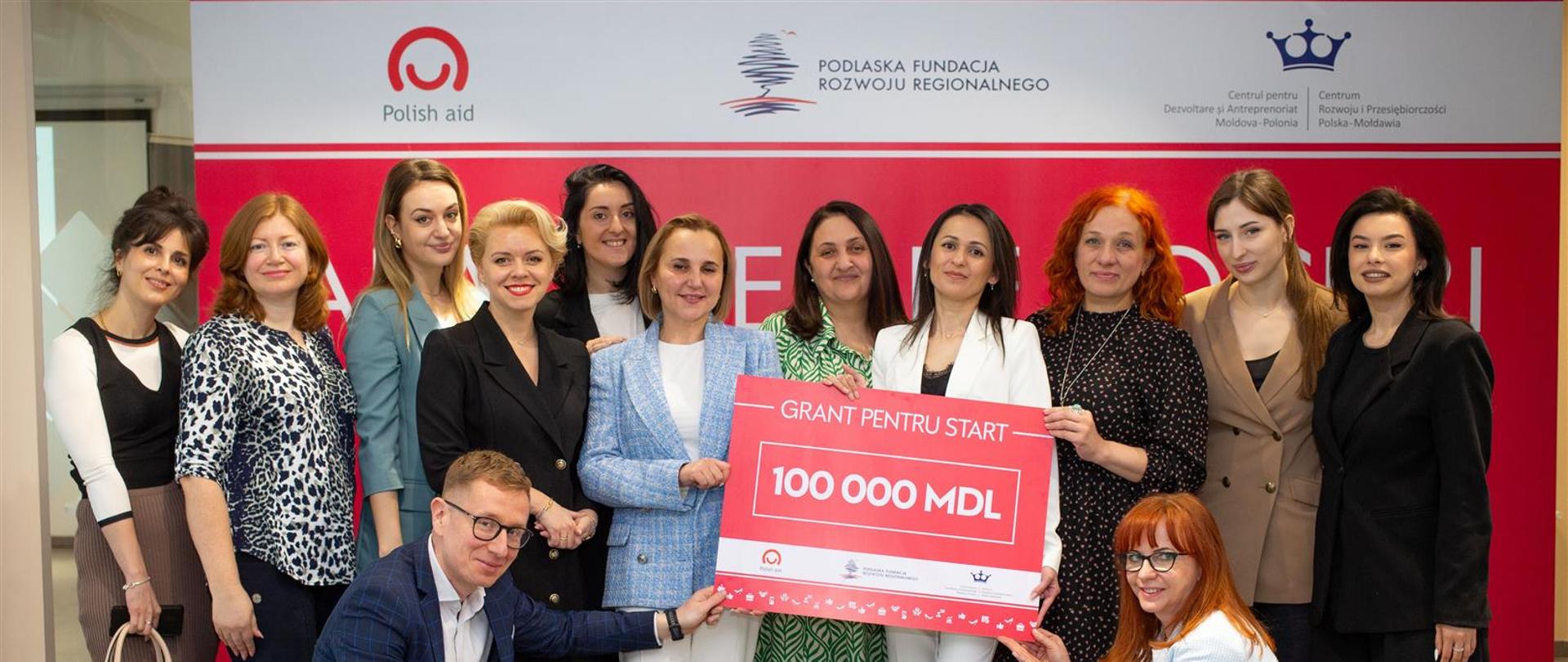Grupa kobiet i 1 mężczyzna pozuje z dużym czekiem na kwotę 100 000 mołdawskich lei. Z tyłu na ściance widać logo Polska Pomoc, Podlaska Fundacja Rozwoju Regionalnego oraz Centrum Rozwoju i Przedsiębiorczości Polska-Mołdawia 