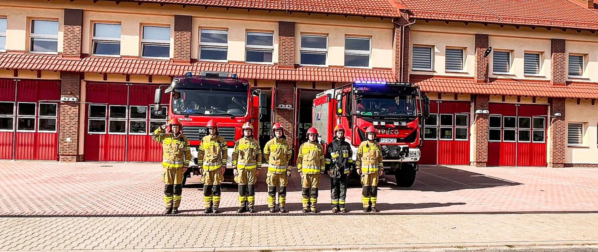 Strażacy w ubraniach specjalnych oddają hołd przy samochodach strażackich przed remizą