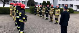 Zdjęcie przedstawia zmianę służbową strażaków na placu apelowym. Na czele szeregu na pierwszym planie stoi oficer ubrany w mundur wyjściowy z czapką rogatywką na głowie. Strażacy stoją frontem do siebie w szeregu dziewięciu na lewej stronie i ośmiu po prawej stronie. Wszyscy strażacy są ubrani w ubrania bojowe typu Moratex, na głowie maja heł, na twarzy maseczki ochronne, na nogach kalosze. Ratownicy po lewo ubrani są w ubrania barwy czarnej z odblaskowymi pasami barwy szarej i żółtej a ratownicy po prawej stronie kadru w ubrania barwy jasno brązowej z odblaskowymi pasami barwy szarej i żółtej. W tle żywopłot oraz budynek Komendy Powiatowej w Łowiczu oraz okna należące do części administracyjnej komendy.