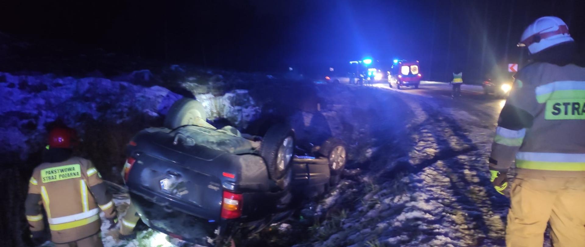 Zdjęcie przedstawia strażaka PSP i OSP oraz samochód osobowy znajdujący się w przydrożnym rowie, na dachu.