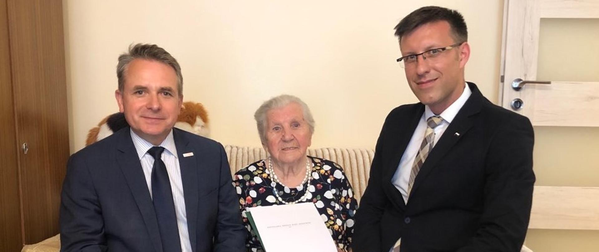 Na zdjęciu znajduje się dwóch mężczyzn i starsza kobieta. Siedzą na kanapie, kobieta między mężczyznami. 