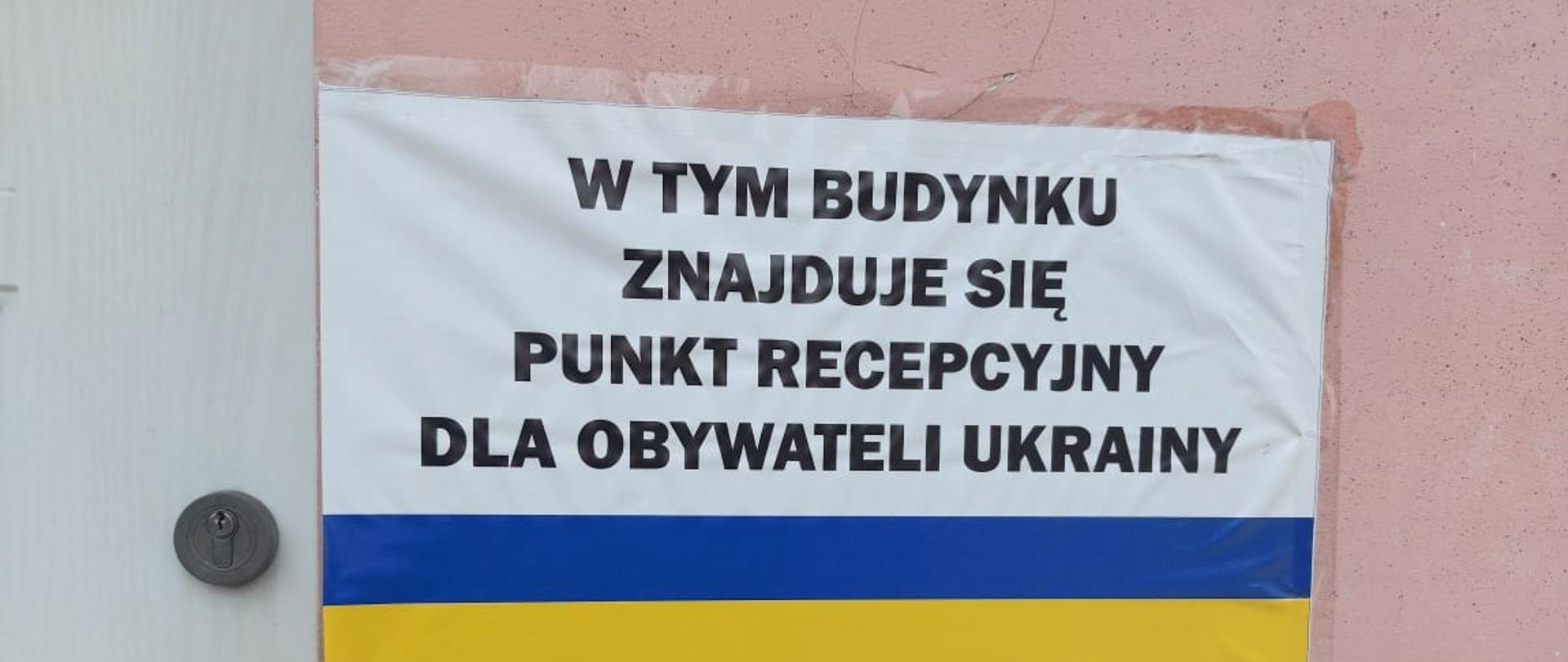 Biała tablica z czarnymi napisami W tym budynku znajduje się punkt recepcyjny dla obywateli Ukrainy, niżej taki sam napis w języku Ukraińskim. Pomiędzy tymi napisami niebiesko-żółty pasek w barwach flagi Ukrainy. Tablica jest przymocowana do różowej ściany budynku. 