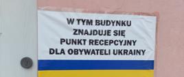 Biała tablica z czarnymi napisami W tym budynku znajduje się punkt recepcyjny dla obywateli Ukrainy, niżej taki sam napis w języku Ukraińskim. Pomiędzy tymi napisami niebiesko-żółty pasek w barwach flagi Ukrainy. Tablica jest przymocowana do różowej ściany budynku. 