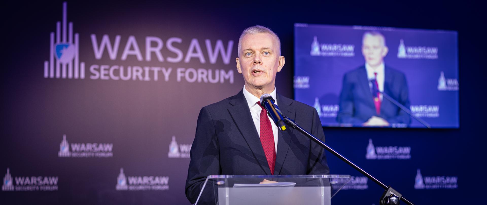 Zdjęcie przedstawia stojącego za mównicą ministra Tomasza Siemoniaka. Z prawej strony mównicy stoi mikrofon na czarnym statywie. W tle ścianka ze zmultiplikowaną nazwą konferencji "Warsaw Security Forum" i logo (białe litery na granatowym tle). Po prawej stronie widać monitor z transmisją obrazu na żywo (przemawiający minister Tomasz Siemoniak).