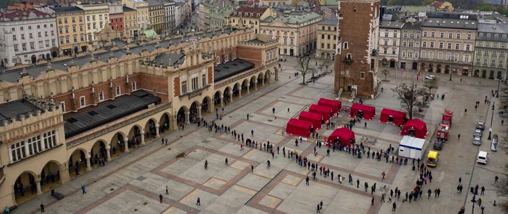 Widok z drona na płytę Rynku Głównego w Krakowie. Na środku czerwone namioty do których oczekuje bardzo długa kolejka osób. W tle wieża Ratusza, po prawej sukiennice.