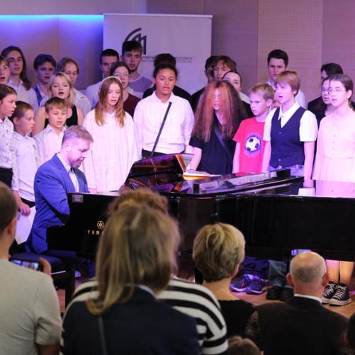 zdjęcie przedstawia chór szkolny wraz z prowadzącym akompaniatorem przy fortepianie na scenie sali koncertowej podczas wykonania utworu, inne ujęcie z bliska