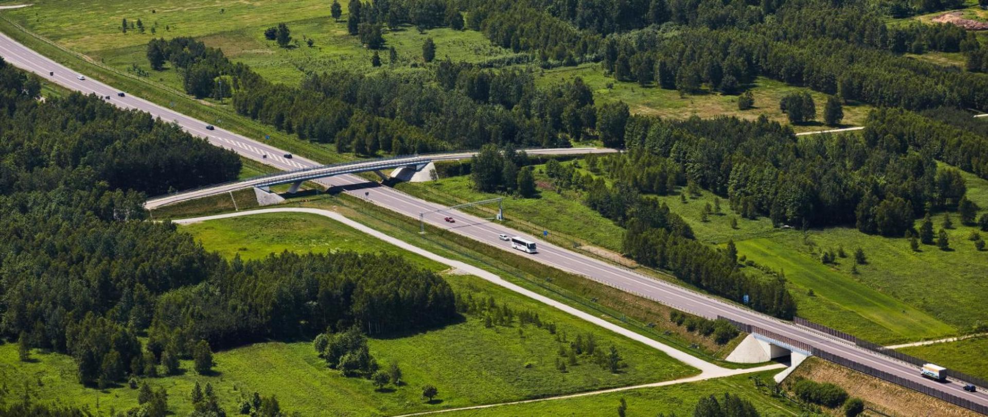 Widok z lotu ptaka na drogę z wiaduktem i przepustem dla zwierząt, otoczoną lasami i zielonymi terenami.