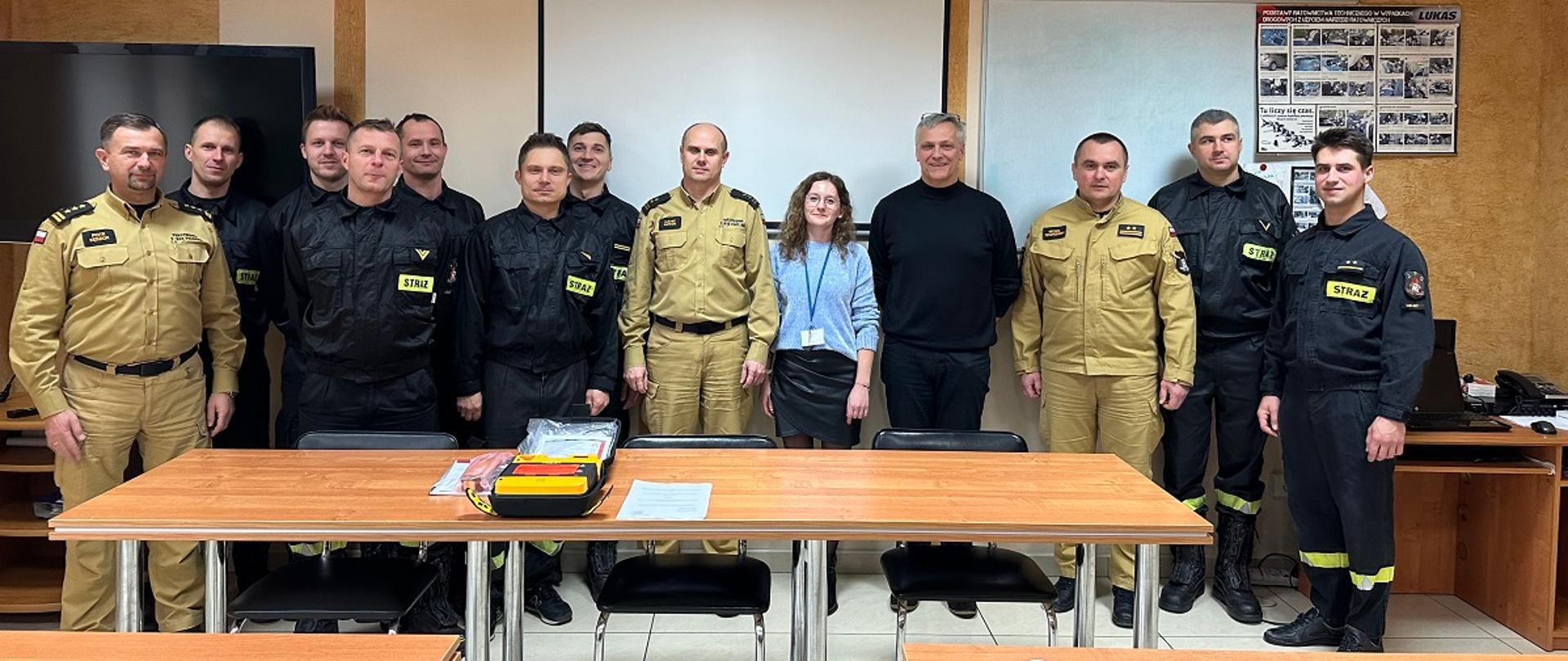 Fotografia wykonana podczas uroczystości przekazania defibrylatora dla mieleckich strażaków. Na zdjęciu stoją funkcjonariusze ubrani w umundurowaniu służbowym oraz przedstawiciele firmy Texen Poland sp. z o.o. Na stole leży podarowany defibrylator.