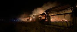 Strażacy prowadzący działania gaśnicze budynku o wymiarach 70m x 25m x 8m, który wypełniony jest balotami słomy oraz składowany jest w nim sprzęt rolniczy. Metalowa konstrukcja dachu przed przybyciem zastępów straży pożarnej uległa zawaleniu do wnętrza budynku. Duże zadymienie, widoczne duże płomienie.