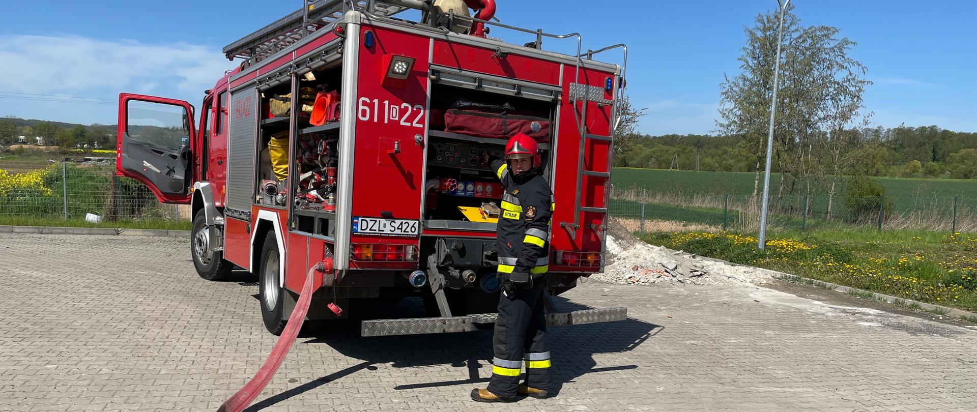 czerwony pojazd pożarniczy, na jego tyle stoi umundurowany strażak, z lewej strony pojazdu widoczny czerwony wąż gaśniczy