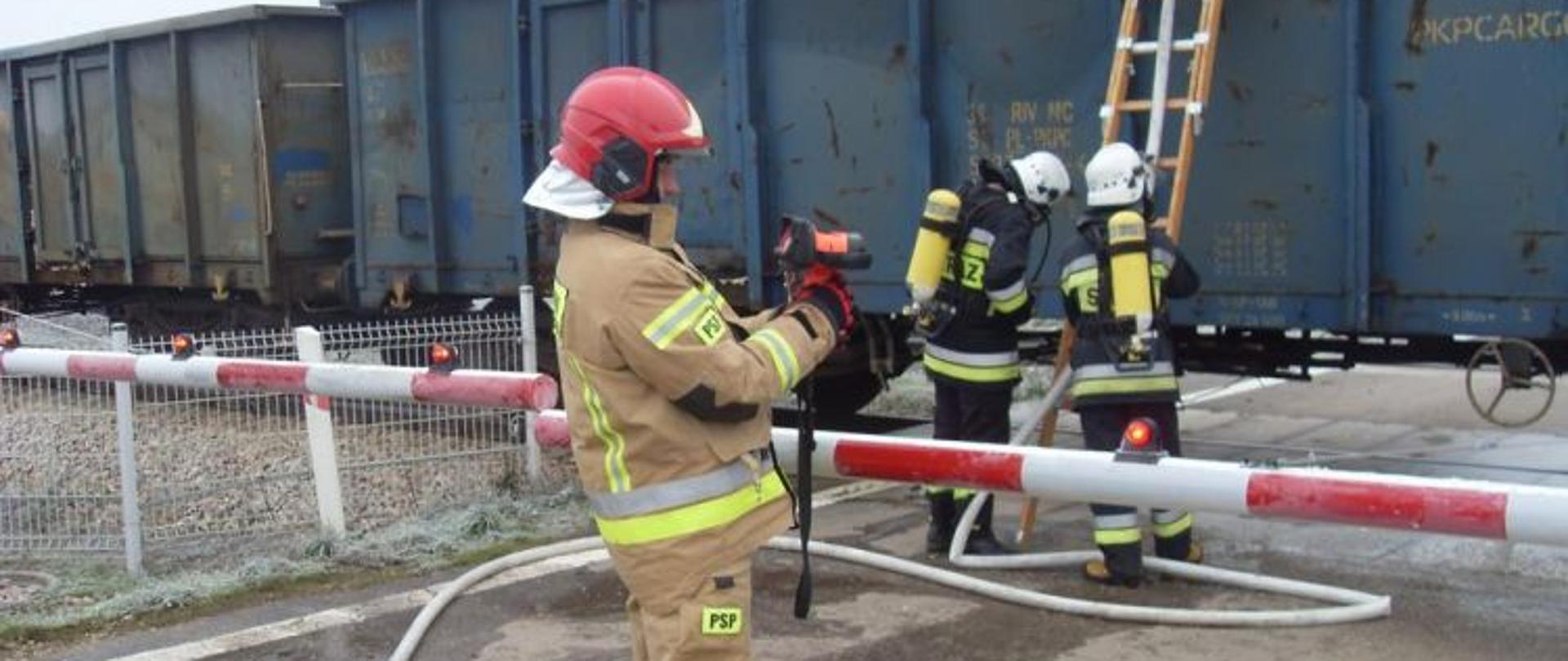 Zdjęcie przedstawia działania straży przy gaszeniu siarki. W tle wagon kolejowy koloru niebieskiego. O wagon oparta drabina nasadkowa na której stoi strażak w pełnym zabezpieczeniu podając prąd wody na ognisko pożaru. Przed drabiną stoi dwóch ratowników zabezpieczając strażaka. Wszyscy maja sprzęt ochrony dróg oddechowych. Na pierwszym planie dowódca w żółtym ubraniu sprawdzający kamera termowizyjną wagon w którym doszło do pożar. Rogatki zamknięte.