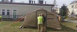 Na zdjęciu widoczny jest rozstawiony namiot ochronny, przeznaczony dla schronienia personelu oraz pacjentów centrum medycznego podczas ćwiczeń. Wokół chodzą strażacy dopinający paski w mocowaniu namiotu.