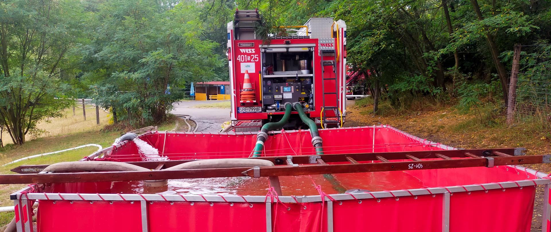 Na zdjęciu widzimy samochód strażacki ustawiony na leśnej drodze. Za pojazdem znajduje się duży, przenośny zbiornik przeciwpożarowy w kolorze czerwonym, wypełniony wodą. Do zbiornika podłączone są węże – jeden doprowadza wodę, a inny zasysa ją do wozu strażackiego.
Zbiornik oparty jest na metalowej konstrukcji i wygląda jak tymczasowy rezerwuar wodny, wykorzystywany podczas akcji gaśniczych w terenie, szczególnie tam, gdzie nie ma hydrantów ani stałego źródła wody. Nad zbiornikiem widoczne są drewniane elementy stabilizujące węże.
Całość znajduje się w otoczeniu zieleni – drzewa i krzewy sugerują, że to teren leśny lub parkowy.