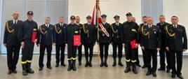 Wspólne zdjęcie grupowe strażaków po uroczystości ślubowania. W centrum poczet sztandarowy z biało-czerwonym sztandarem, a po obu stronach stoją strażacy w mundurach galowych i służbowych, niektórzy trzymają czerwone teczki z aktem ślubowania.