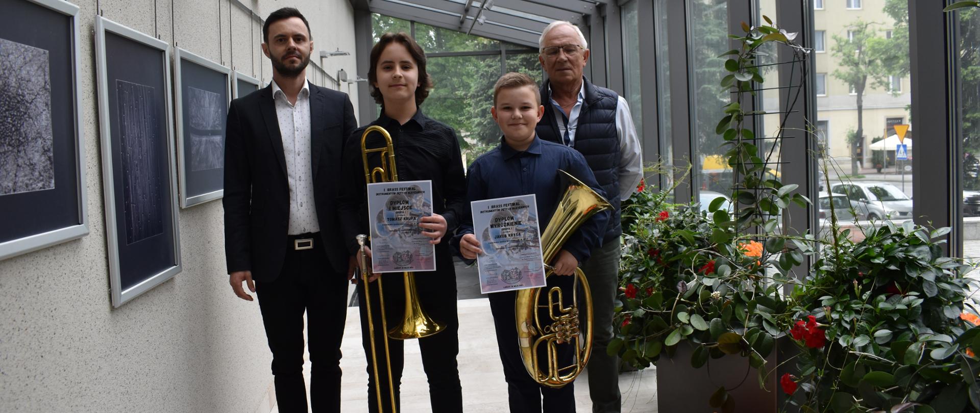Na zdjęciu w centralnej części stoją laureaci konkursu prezentujący instrumenty i dyplomy wraz z nauczycielem instrumentu głównego i akompaniatorem. Z lewej strony na tle jasnej ściany zawieszone są współczesne grafiki w kolorach czarno-szarym, po prawej stronie na tle dużych okien widnieje kwiatowa ekspozycja.