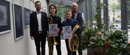 Na zdjęciu w centralnej części stoją laureaci konkursu prezentujący instrumenty i dyplomy wraz z nauczycielem instrumentu głównego i akompaniatorem. Z lewej strony na tle jasnej ściany zawieszone są współczesne grafiki w kolorach czarno-szarym, po prawej stronie na tle dużych okien widnieje kwiatowa ekspozycja.