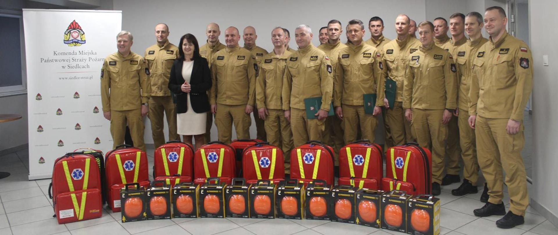 Przekazanie plecaków ratowniczych. W tle funkcjonariusze PSP wraz z Komendantem Wojewódzkim i panią Marszałek Województwa, przekazujący plecaki ratownicze