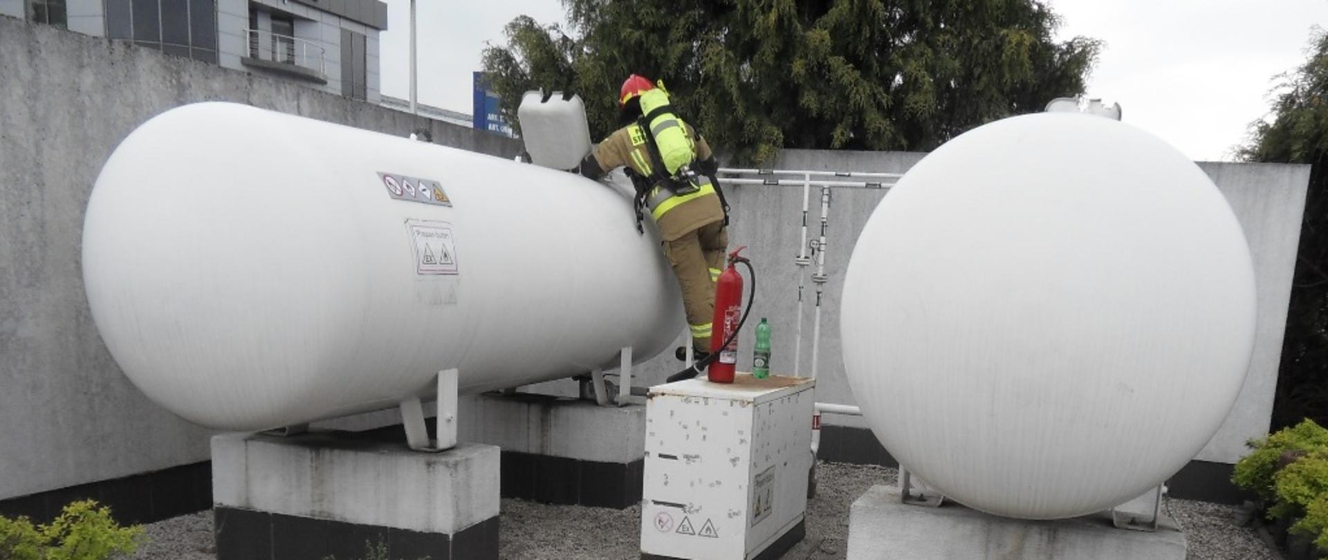 Na zdjęciu dwa zbiorniki z gazem LPG, w jednym ze zbiorników strażak uszczelniający uszkodzony manometr przy użyciu zwilżonej tkaniny i gaśnicy śniegowej