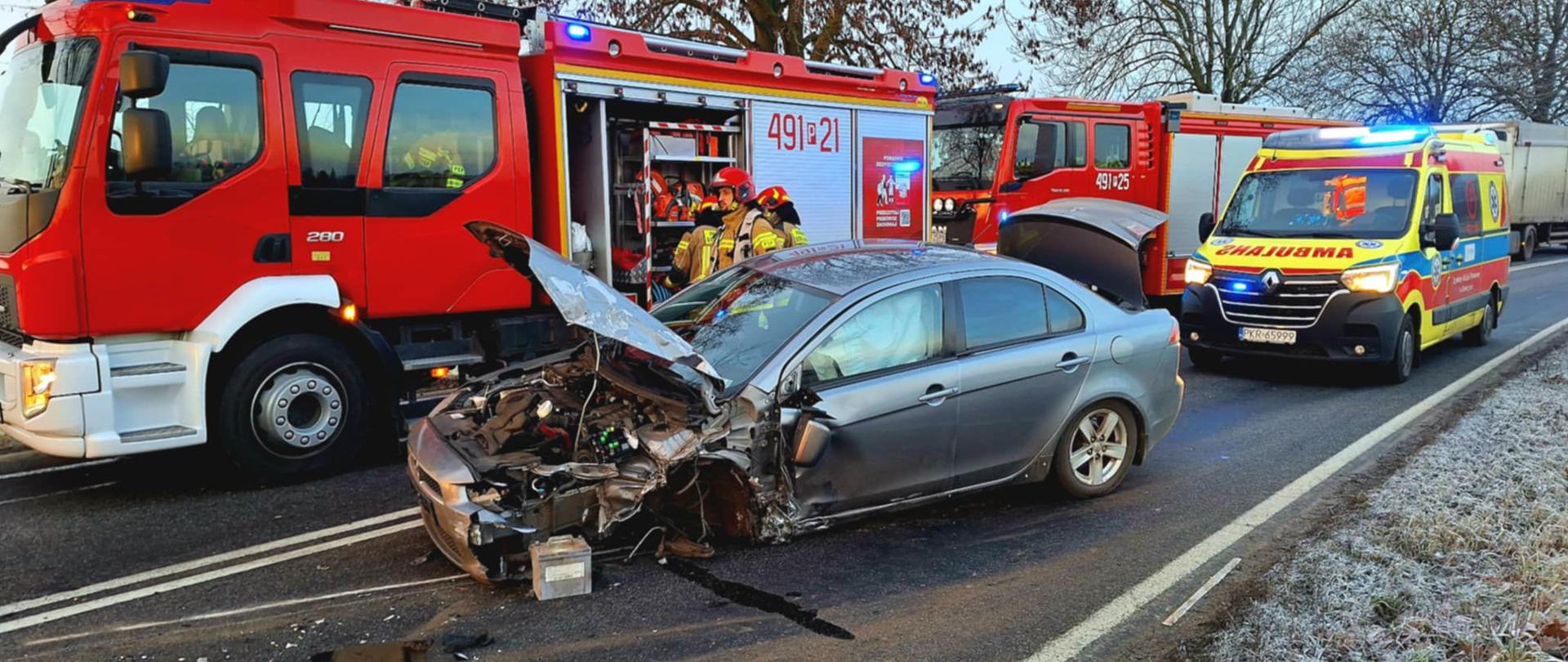 Zdjęcie przedstawia uszkodzony samochód osobowy na drodze, a zanim wozy strażackie oraz samochód zespołu ratownictwa medycznego.