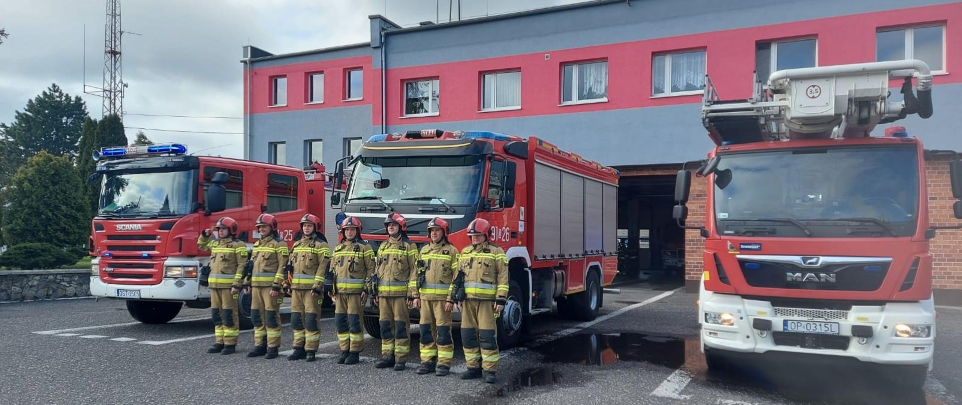 Na zdjęciu znajdują się strażacy ustawieni na tle samochodów strażackich wystawionych przed garaż