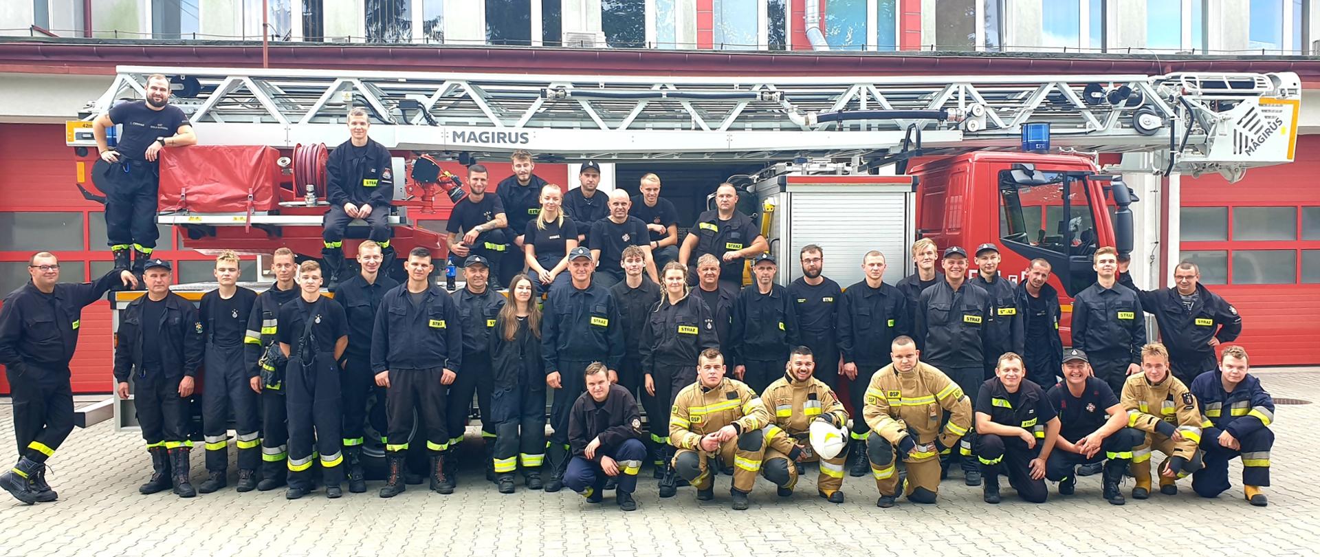 Grupa strażaków w ubraniach koszarowych i bojowych na tle samochodu z drabiną pożarniczą. W głębi budynek komendy.