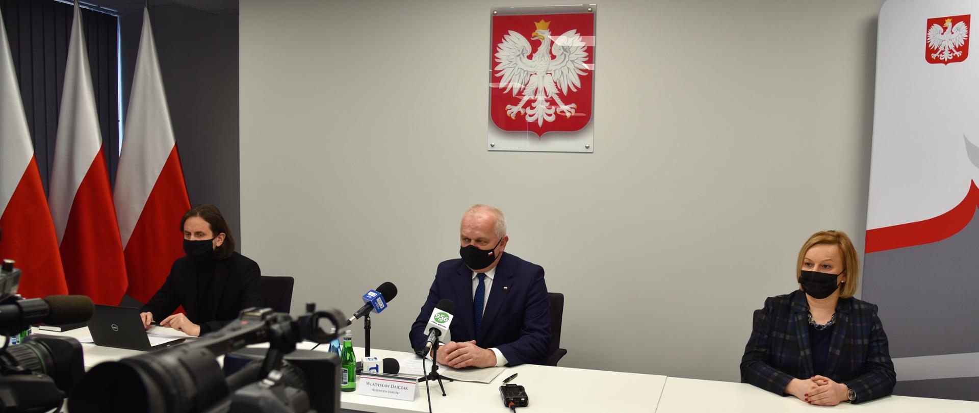 Na pierwszym planie dwie kamery telewizyjne. W części centralnej długi, biały stół konferencyjny. Za nim siedzą, w oddaleniu od siebie (od lewej) dyrektor NFZ-u, wojewoda lubuski i burmistrz Drezdenka. W tle roll-up Lubuskiego Urzędu Wojewódzkiego i biało-czerwone flagi