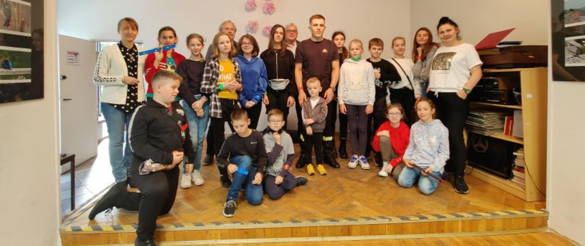 Zdjęcie grupowe wykonane wewnątrz Klubu Osiedlowego. Na zdjęciu w szeregu stoją dzieci i opiekunowie. Na środku strażak w ubraniu koszarowym