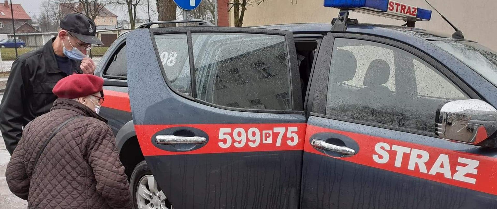 Widać strażaka, który pomaga starszej kobiecie wejść do samochodu straży pożarnej
