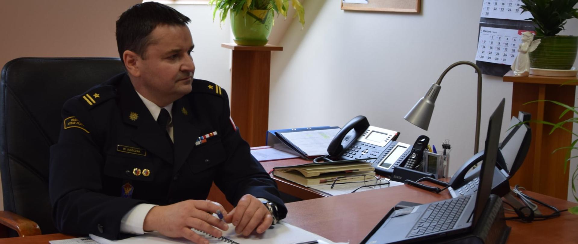 Strażak w umundurowaniu galowym siedzi przy biurku, w dłoni trzyma długopis, jego wzrok jest skierowany na komputer przenośny.