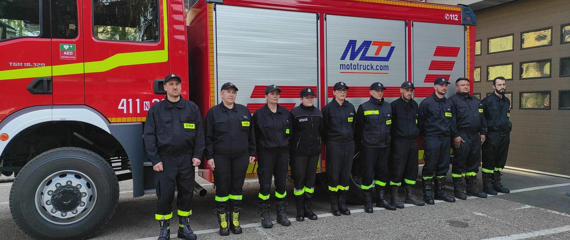 Na tle dużego samochodu strażackiego stoi grupa strażaków ochotników w czarnych mundurach