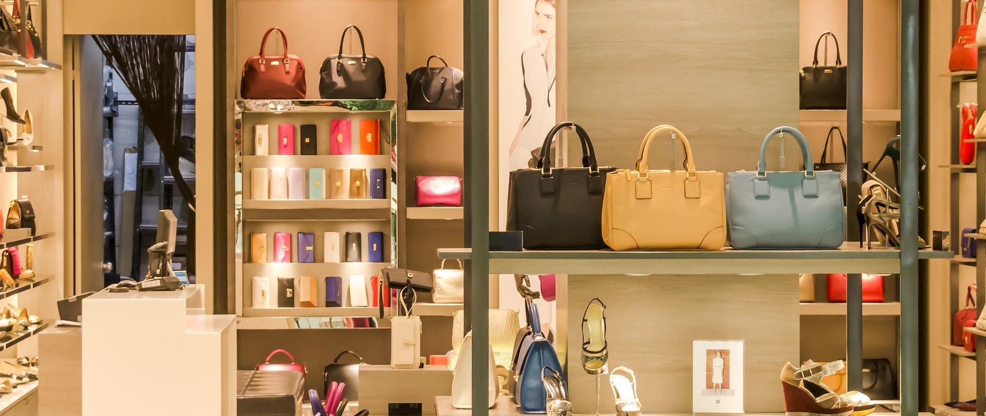 Zdjęcie przedstawia sklep z budai i akcesoriami. Na pierwszym planie jest półka z torebkami, na dalszym portfele i buty.