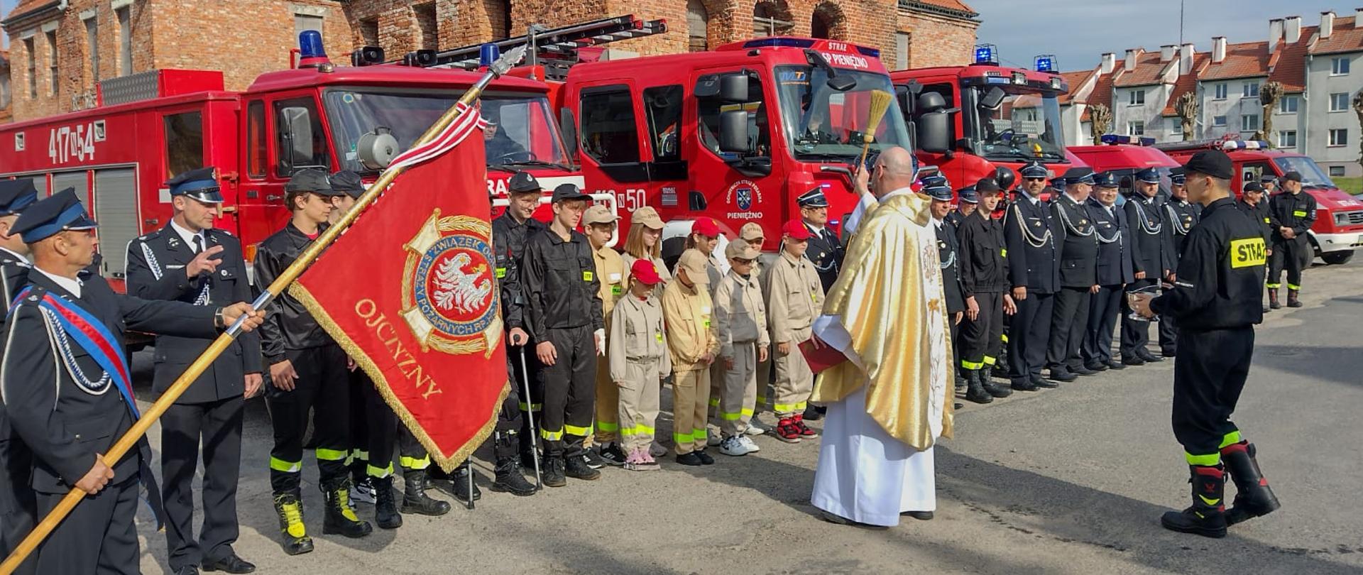 Strażacy ochotnicy w mundurach galowych, dzieci z drużyny pożarniczej. Z lewej strony sztandar. W tle samochody strażackie. Ksiądz z kropidłem święci samochody strażackie.