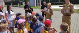Strażak pokazujący czujki, przed nim zgromadzone dzieci na placu komendy