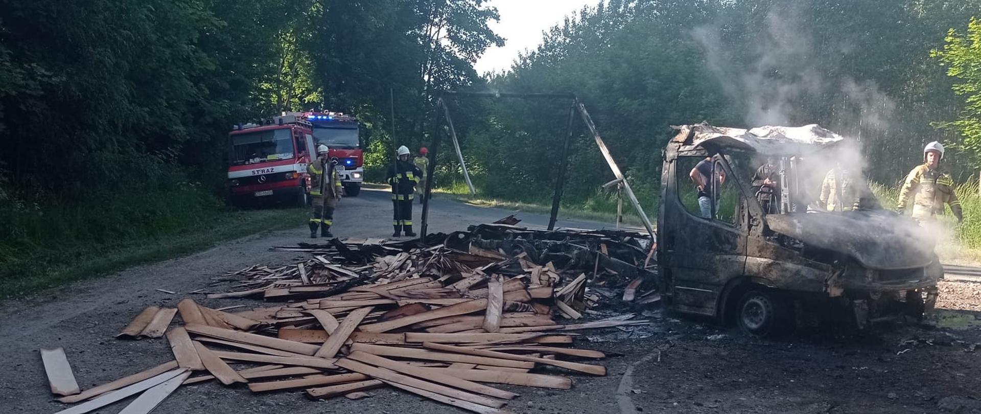 Zdjęcie przedstawia wrak auta dostawczego po spaleniu, który znajduje się na poboczu drogi .Obok auta znajdują się strażacy oraz wozy strażackie.
