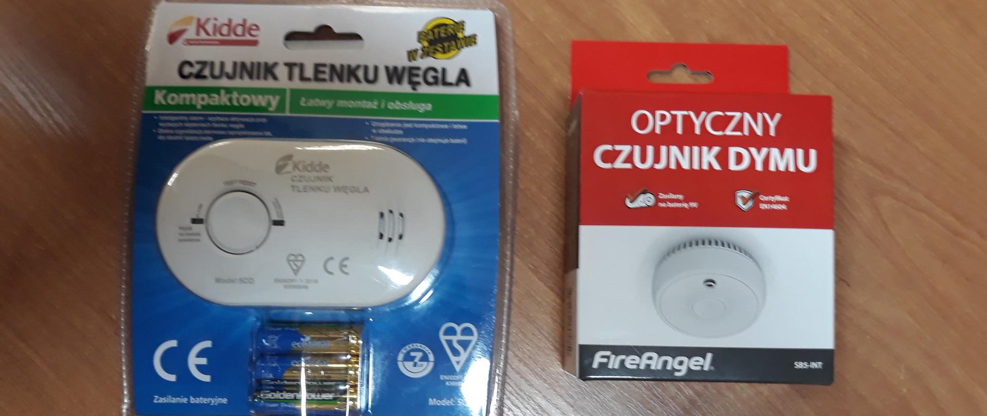 Na blacie stołu leży fabrycznie zapakowany w plastikowy blister czujnik tlenku węgla, wraz z kompletem baterii, które go zasilają obok niego w kartonowym opakowani w kolorach czerwonym białym i czarnym znajduje się optyczny czujnik dymu.