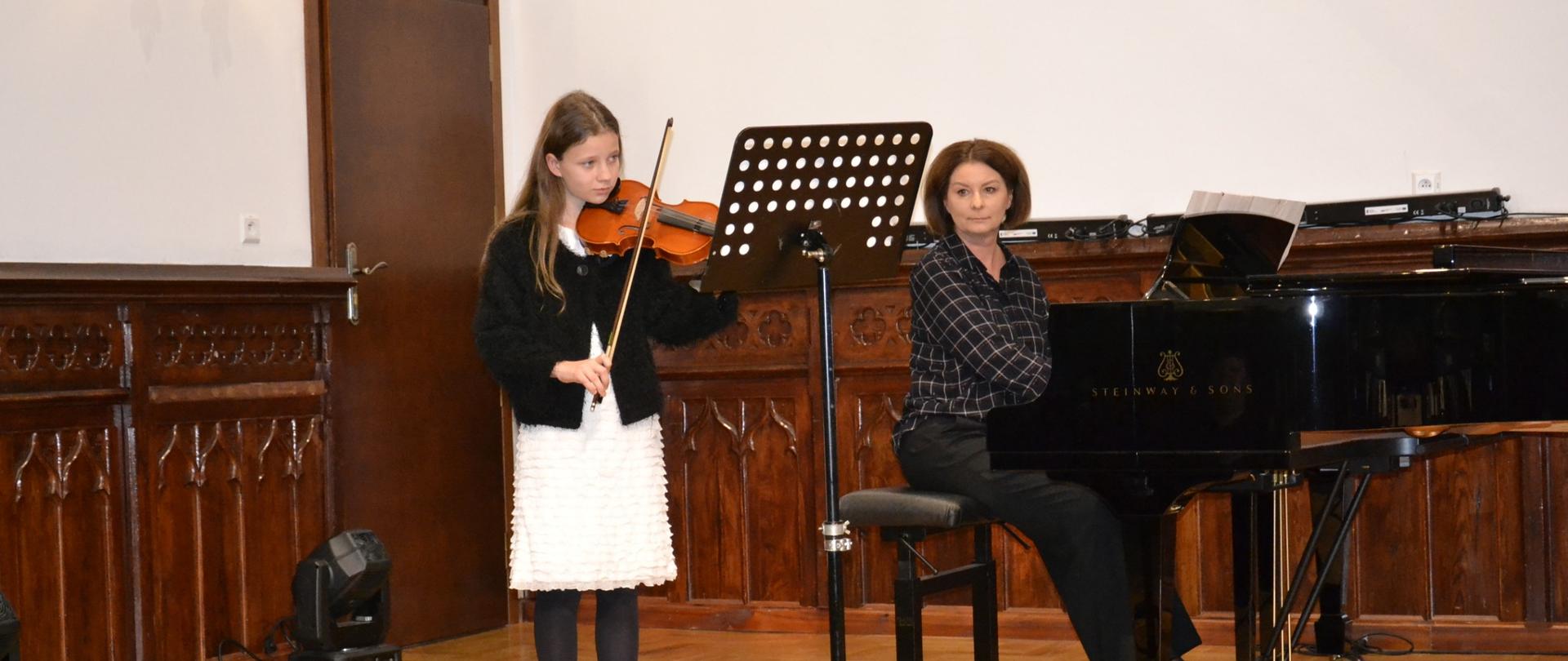 Dziewczynka grająca na skrzypcach podczas koncertu klasowego, akompaniowana przez nauczycielkę przy fortepianie na scenie szkolnej.